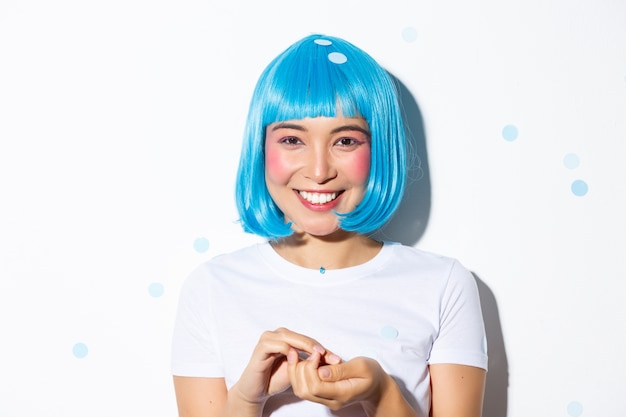 Крупный план милой азиатской девушки в синем парике, празднующей Хэллоуин, бросая конфетти и улыбаясь, стоя.