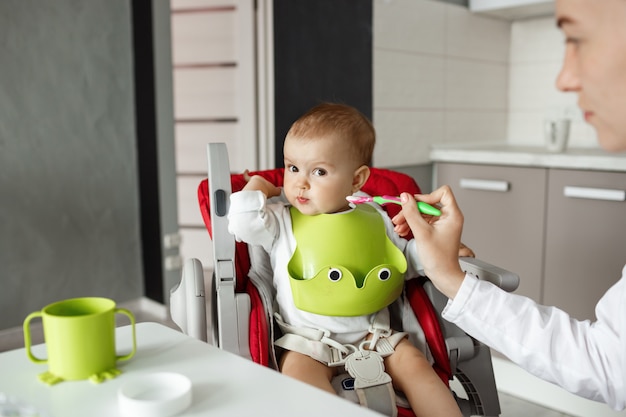 Close up of cute baby son sitting in kitchen in baby chair and turning head aside refusing to eat baby food. Mother tries to feed him with spoon.