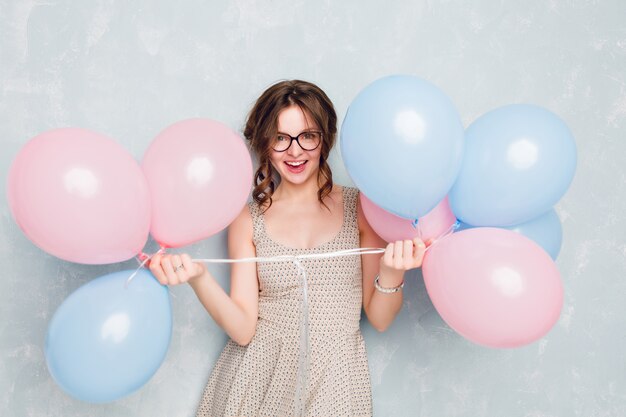 Close-up of cute brunette girl standing in a studio, smiling widely and playing with blue and pink balloons. She is having fun