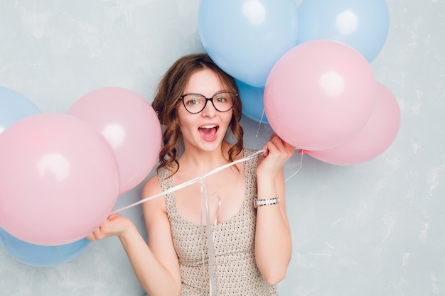 Close-up of cute brunette girl standing in a studio, smiling widely and playing with blue and pink balloons. She is having fun