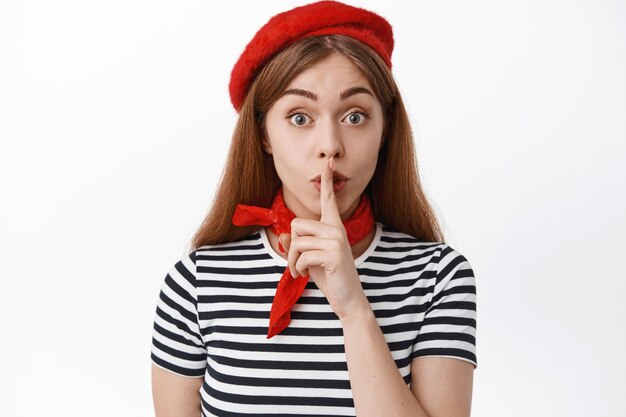 Close up of cute female model in french red beret hiding a secret, making shh shush sign, press finger to lips, ask to be quiet, stay silent, standing over white wall