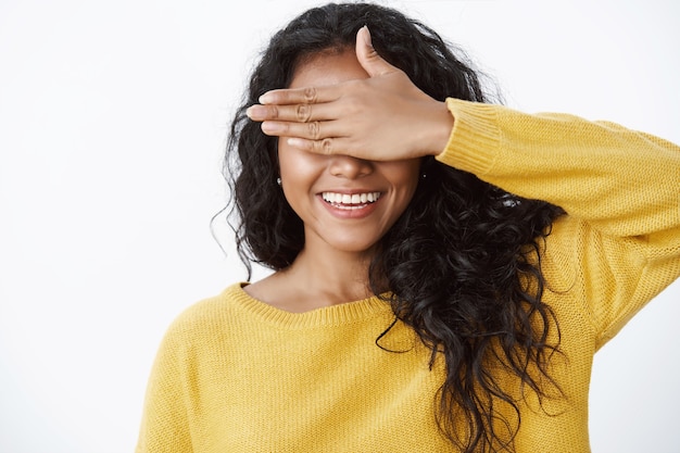 Foto gratuita primo piano carino donna dai capelli ricci femminile in maglione giallo copre gli occhi con il palmo e sorride felicemente, aspettando la sorpresa di compleanno, giocando a nascondino, aspettandosi qualcosa