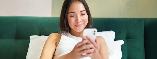 Close up of cute girl lying in bed looking at smartphone reading on mobile phone messaging