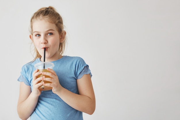 Close up of cute girl with light hair in tail hairstyle holding glass of milk in hands, drinking through straw and looking aside with funny face.