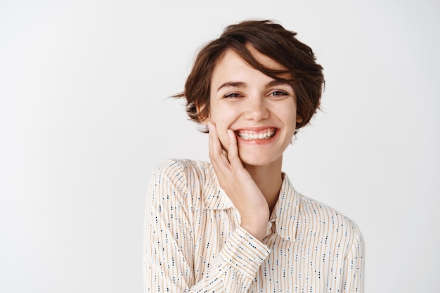 Close up of cute girl with short hair smiling with white teeth and touching natural clean face, standing over wall