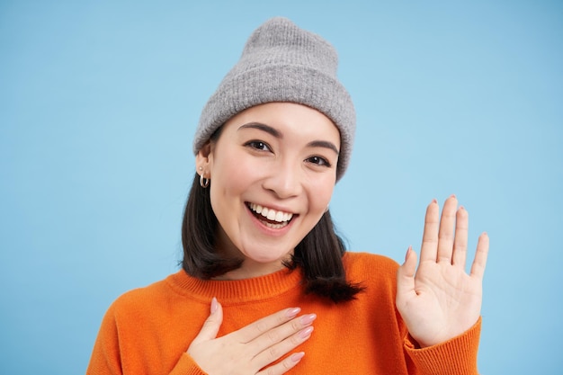 Free photo close up of cute korean girl in beanie raises one hand and introduces herself greets you smiles and