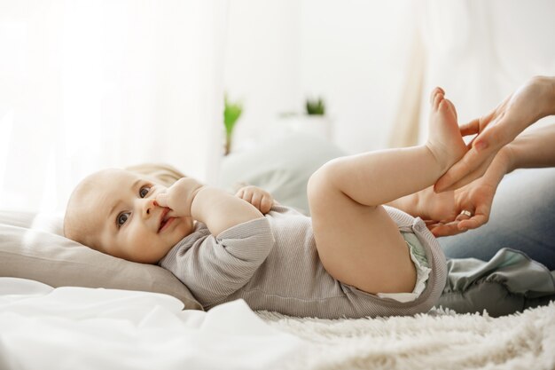 Close up of cute newborn baby lying on bed, looking aside while mother playing and touching his little legs. Baby gnawing finger, his face express happiness and joy.