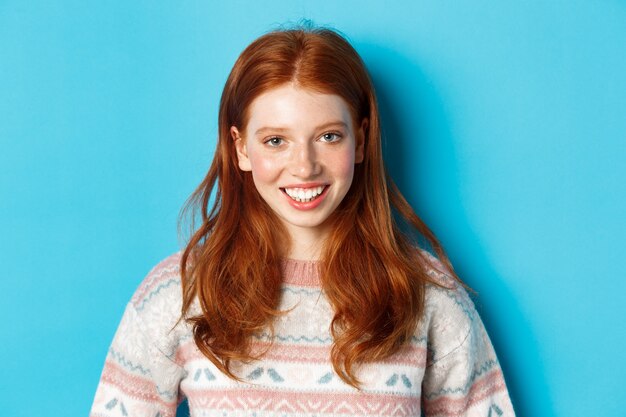 青い背景に立って、カメラで幸せそうに笑っているセーターのかわいい赤毛の女の子のクローズアップ。