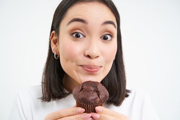 Free photo close up of cute smiling asian woman holding chocolate cupcake near mouth having bite enjoys eating