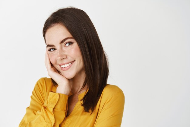 Close-up of cute smiling woman looking flirty at camera, touching cheek and blushing, standing on white wall in yellow shirt