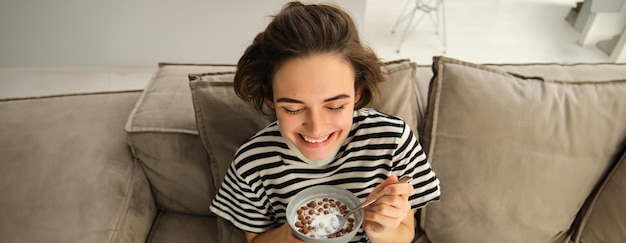 Free photo close up of cute young female model eating cereals with milk enjoys her breakfast on sofa in living