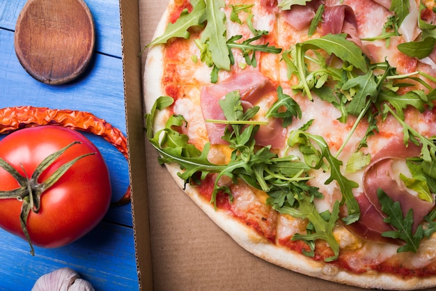 Close-up of delicious bacon and arugula pizza in box with garlic; tomato and chili on table