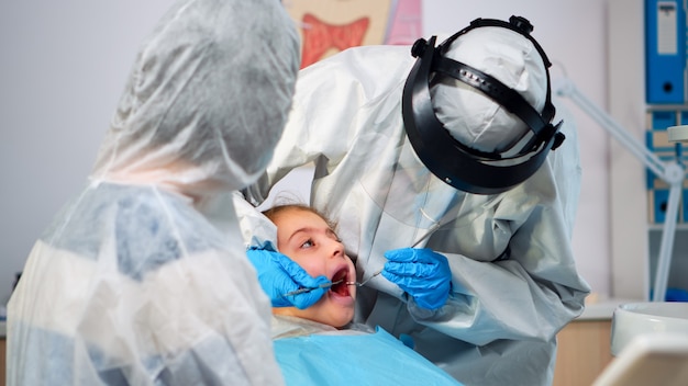 Закройте стоматолога в комбинезоне, исследующего пациентку в новом нормальном стоматологическом кабинете, стоя возле стоматологического кресла. Концепция посещения врача во время вспышки коронавируса с защитной маской