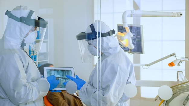 Close up of dentist doctors with face shield and ppe suit discussing in dental room about teeth digital x-ray while patient waiting. Concept of new normal dentist visit in coronavirus outbreak.