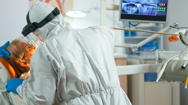 Close up of dentistry doctor in coverall using drilling machine for examining patient during global pandemic. Medical team wearing protective suit,, face shield, mask, gloves in stomatological office