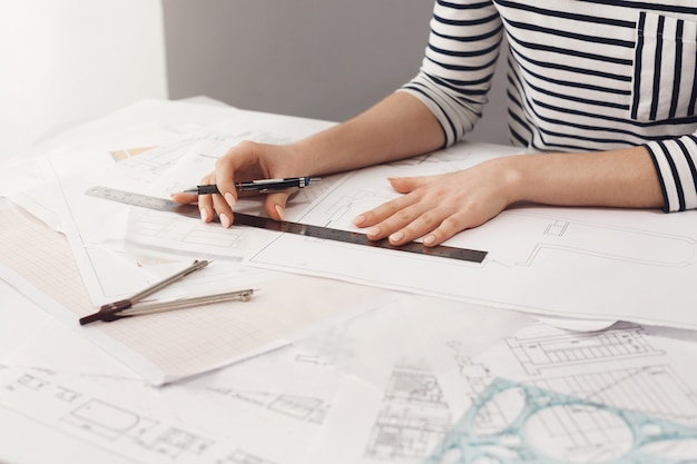 Free photo close up detail of beautiful young female architect in striped shirt sitting at desk, holding pen and ruler in hands, making blueprints, working on new project at home. business and art