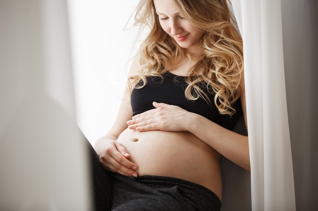 Закройте вверх по детали молодой белокурой беременной матери в черном обмундировании сидя на подоконнике в спальне, касаясь и смотря животу с счастливым выражением.