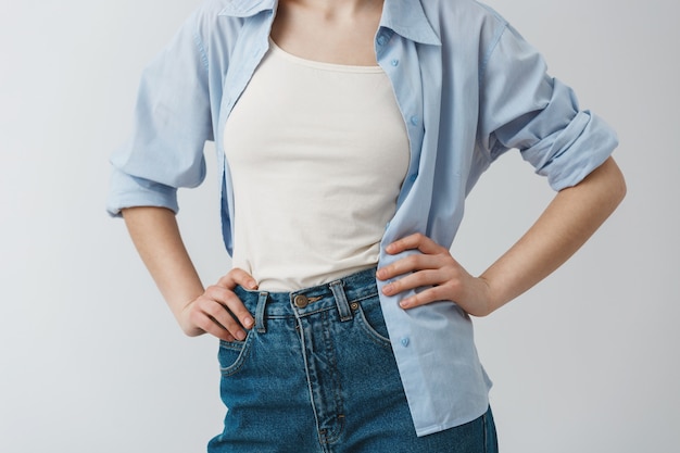 Close up detail of stylish clothes of young female student holding hands on waist, wearing white t-shirt under blue shirt and jeans.