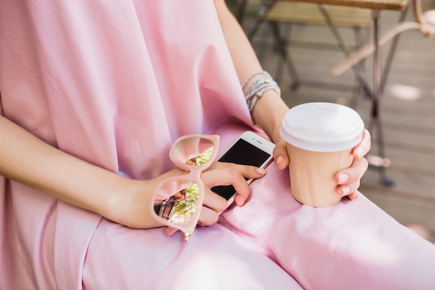 Chiudere i dettagli delle mani della donna seduta al caffè in abito di moda estiva, stile hipster, abito di cotone rosa, occhiali da sole, bere caffè, accessori eleganti, abbigliamento rilassante e alla moda