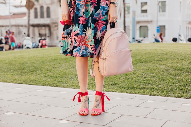 Chiudere i dettagli delle gambe in sandali rosa della donna alla moda che cammina nella strada della città in gonna colorata stampata, tenendo lo zaino in pelle rosa