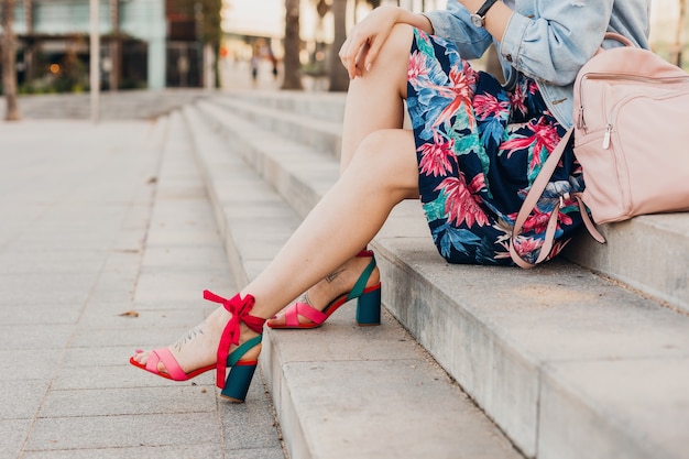 Close up details of legs in pink sandals of woman sitting on stairs in city street in stylish printed skirt with leather backpack, summer style trend