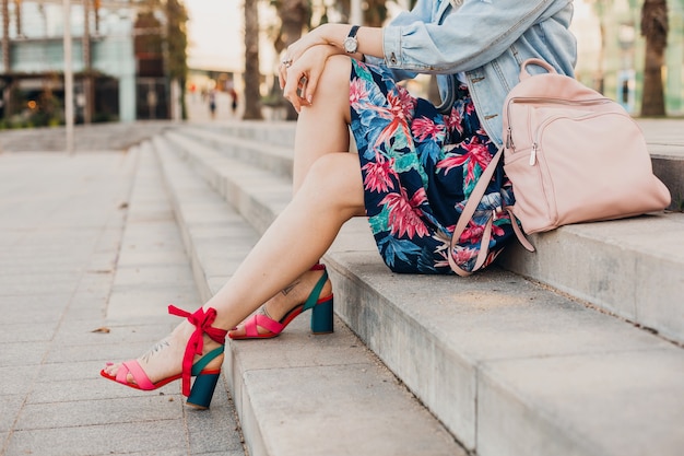 革のバックパック、夏のスタイルのトレンドとスタイリッシュなプリントスカートの街の通りの階段に座っている女性のピンクのサンダルの足の詳細を閉じる