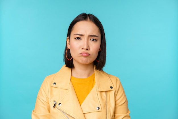 Close up of disappointed asian girl sulking grimacing and frowning displeased standing moody against blue background Copy space