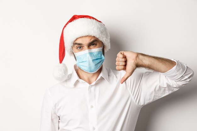  Close-up of disappointed man in face mask and christmas hat showing thumb down, standing  
