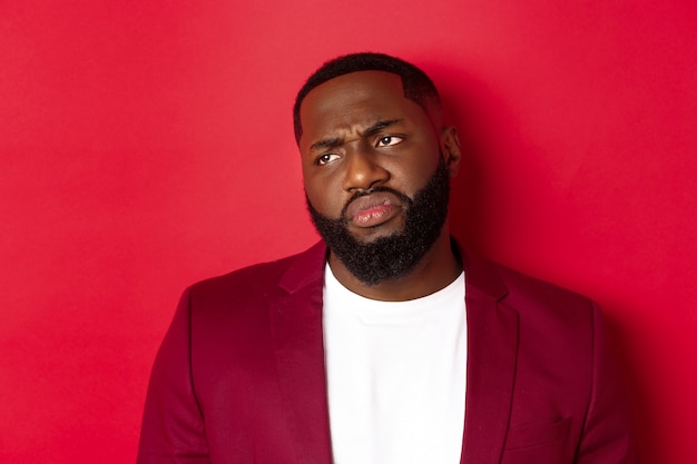 Close-up of disappointed and skeptical Black man looking left, grimacing and sulking displeased, standing over red background