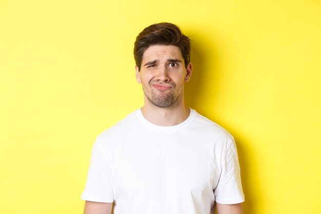 Close-up of dispelased young man in white t-shirt looking doubtful, grimacing unsatisfied, standing over yellow background.