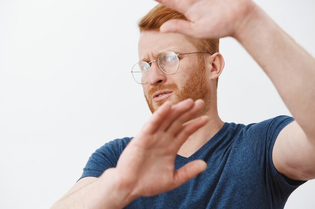 Close-up of displeased adult man in glasses covering his face with hands from light, defending himself