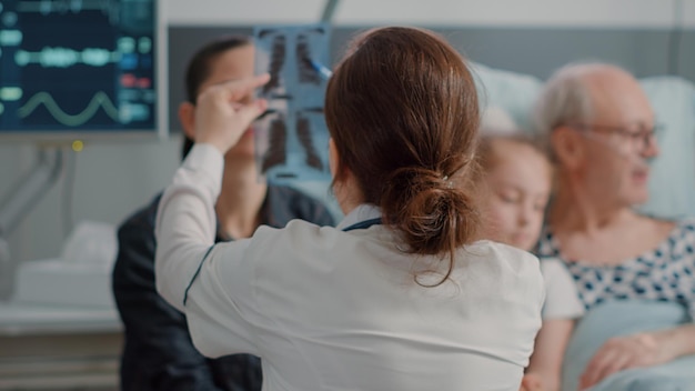 Close up of doctor showing x ray scan to daughter of patient in hospital ward. General practitioner presenting radiography diagnosis to woman while child visiting elder man at intensive care.