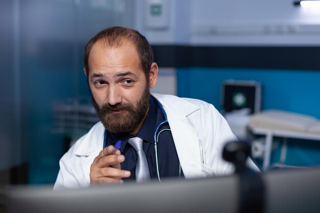 患者とのビデオ通話のためにコンピューターでウェブカメラを使用している医師のクローズアップ。深夜に働いて、遠隔検診の予約のためにオンライン会議で話している医者として働いている男。