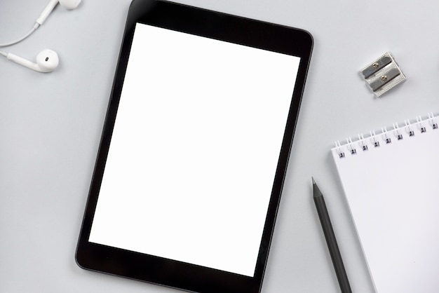 Close-up of earphone; blank digital tablet; sharpener; pencil and spiral notepad on gray background