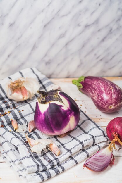Close-up of eggplants; onion; garlic cloves and chequered pattern textile on wooden surface
