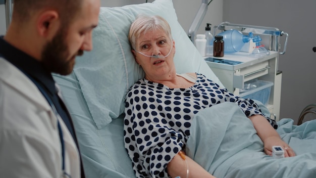 Close up of elder patient laying in bed and talking to medic for healthcare treatment. Doctor consulting sick woman with nasal oxygen tube and IV drip bag for recovery in hospital ward