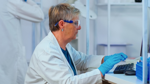 Close up of elderly chemist woman typing on computer in modern equipped laboratory. Multiethnic stuff analysing vaccine evolution using high tech for researching treatment against covid19 virus