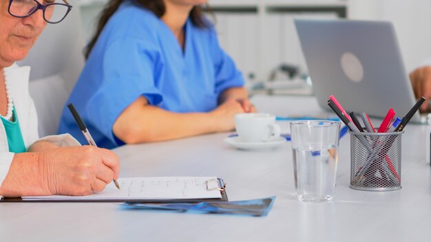 Close up of elderly woman doctor taking notes on clipboard while coworkers discussing in background, writing on laptop during brainstorming. Profesional teamworker having medical meeting