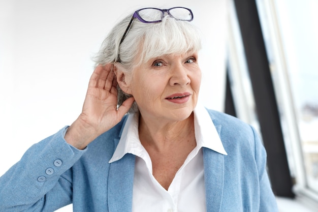 Close up elegant stylish elderly woman in formal suit having hearing problems, holding hand at her ear, trying to hear you, saying: Speak louder, please. Age, maturity, people and health concept