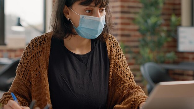 Close up of employee working with laptop and taking notes on papers with charts, wearing face mask. Woman using computer to work on business project during coronavirus pandemic.