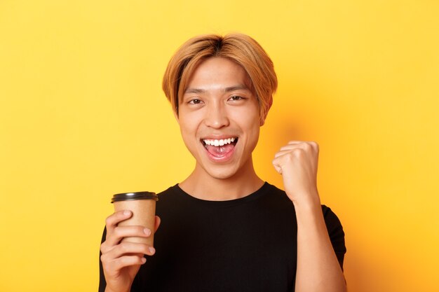 Close-up of energized handsome asian guy fist pump joyfully while drinking coffee, smiling excited over yellow wall.