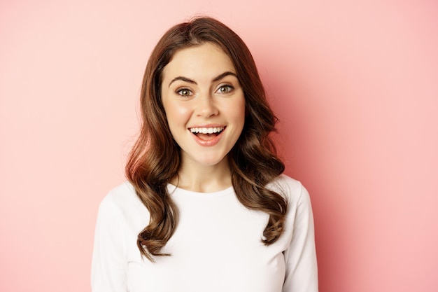 Close up of enthusiastic brunette girl with makeup, smiling and looking happy at camera, posing against pink background.
