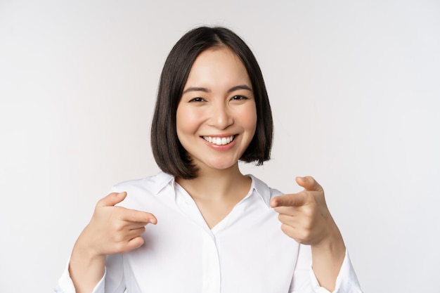 Close up of enthusiastic young woman smiling pointing fingers at camera choosing you inviting and recruiting people standing over white background