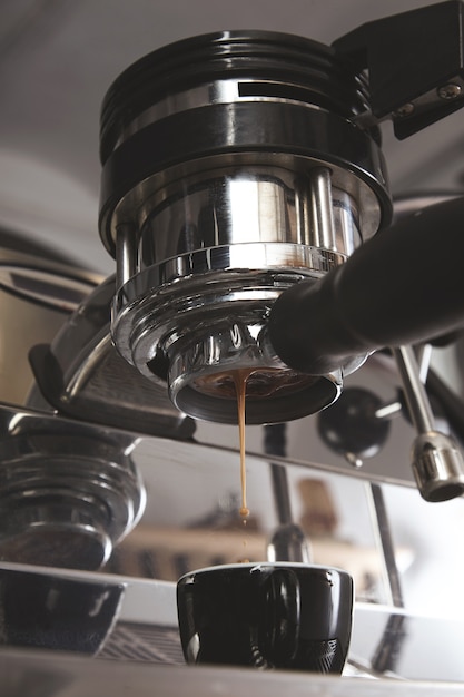 Close up of espresso pouring from silver metallic coffee machine in black ceramic cup. Professional coffee brewing. Drops of roasted coffee fall from top.