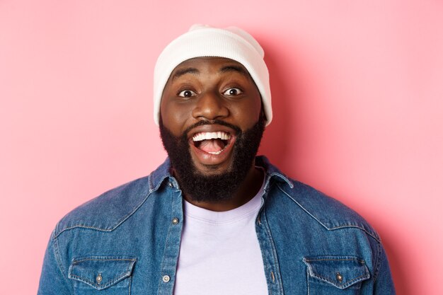 Close-up of excited bearded african-american guy in beanie staring at camera, express amazement and joy, standing over pink background