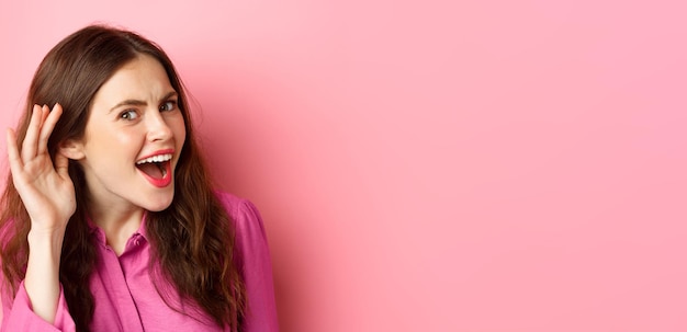 Close up of excited young woman overhear juice rumor lean closer to camera with hand near ear eavesd