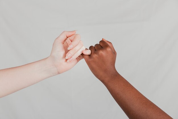 Close-up of fair and dark women hands making a pinkie promise against grey backdrop