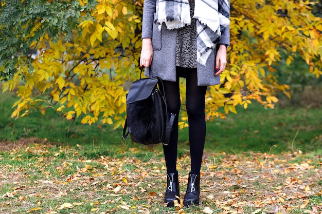 Close up fashion details, woman posing at city autumn park, street style look, trendy leather boots, backpack, luxury dress and coat, bright colors.