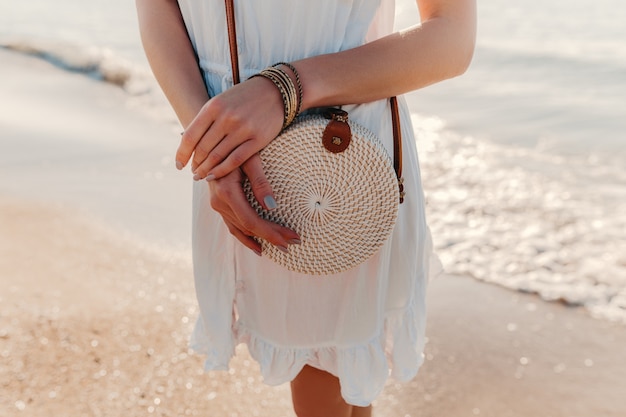Close-up fashion details of woman in white dress with straw purse bag summer style on beach accessories