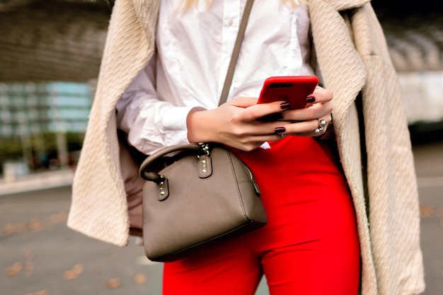 Close up fashion details you business woman , tapped something on her phone, urban autumn city background, bright suit and cashmere coat, ready for conference.